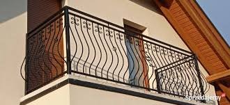 Czarna, metalowa balustrada balkonowa z dekoracyjnymi, falistymi prętami, zamontowana na jasnej elewacji budynku.