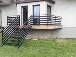 Zewnętrzne schody metalowe z czarną, poziomą balustradą prowadzące do jasnego wejścia domu, widoczne trawnik i fragment elewacji.