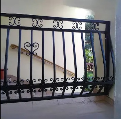 Czarna, ozdobna barierka metalowa z motywem serca na tle jasnych schodów z drewnianą poręczą i widokiem na zieleń ogrodu.