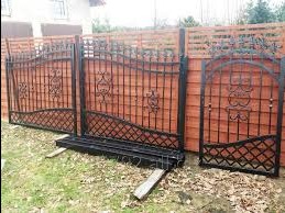 Elementy kutego ogrodzenia w kolorze czarnym, oparte o drewniane panele ogrodzeniowe, z widocznym fragmentem trawnika i domu w tle.