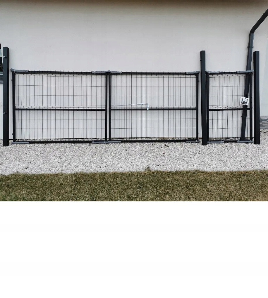 Panelowe ogrodzenie z czarnej siatki metalowej na tle jasnej ściany budynku, ułożone na żwirowym podłożu, z widocznym fragmentem trawnika.