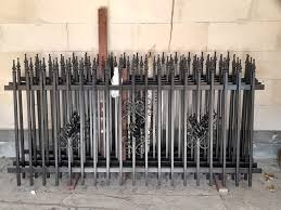 Elementy kutego ogrodzenia w kolorze czarnym, ułożone poziomo na podłodze w surowym pomieszczeniu z ceglanymi ścianami, oczekujące na montaż.