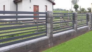 Nowoczesne ogrodzenie posesji z poziomych, szarych profili metalowych, osadzone na niskim murku z betonowych bloczków, widok z zewnątrz, w tle budynek mieszkalny i zieleń.