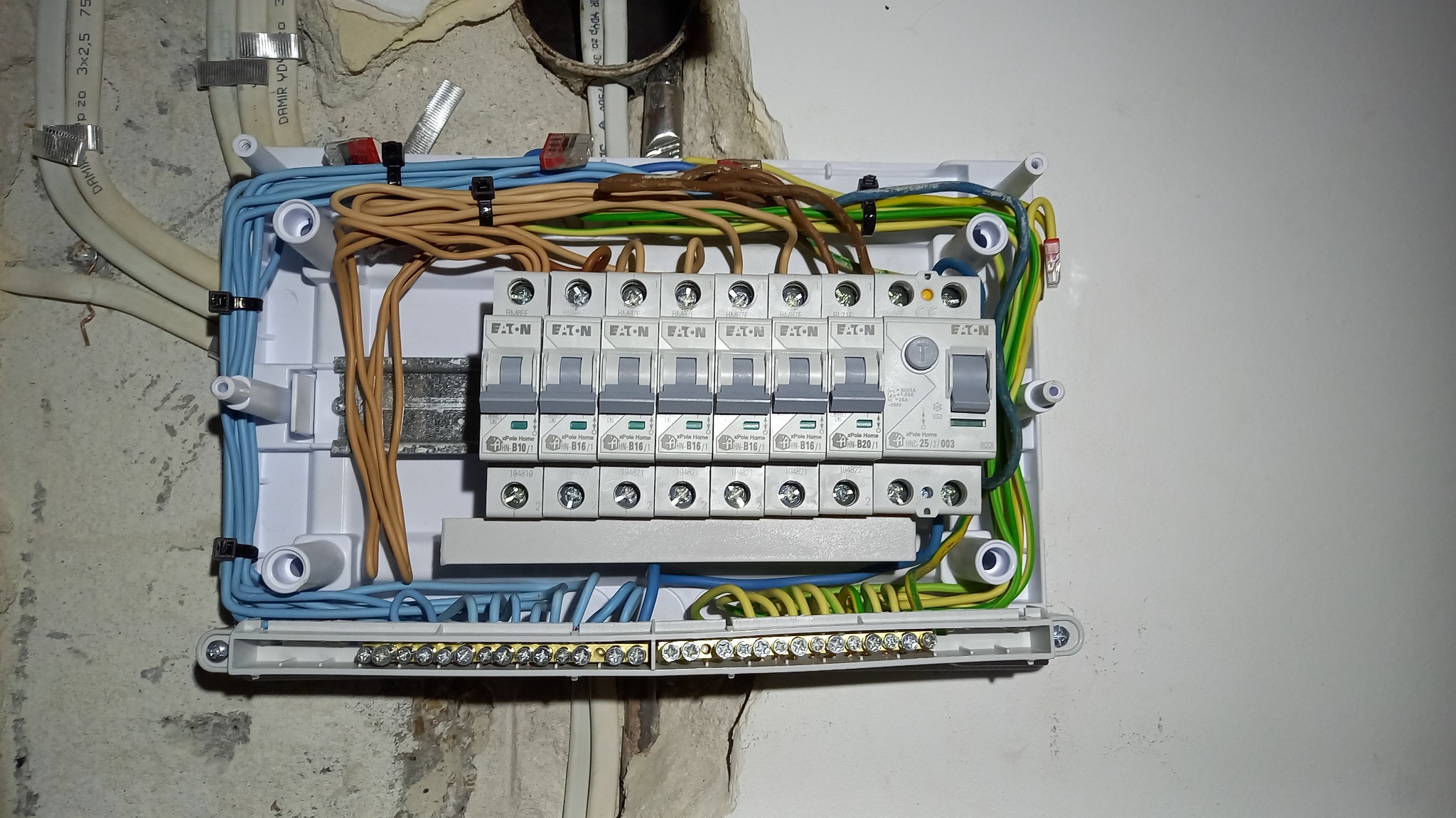 Otwarta skrzynka rozdzielcza z zainstalowanymi wyłącznikami nadprądowymi Eaton, widoczne przewody w różnych kolorach podłączone do listwy zaciskowej i wyłączników, zamocowana na szarej ścianie.