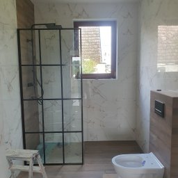 Grzegorz Szczupacki - Nowoczesna łazienka z kabiną prysznicową typu walk-in z czarnymi profilami, marmurowymi płytkami na ścianach, drewnopodobną zabudową WC i wiszącą miską ustępową, widoczna drabina malarska.