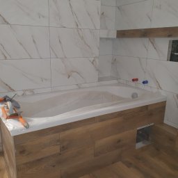 Grzegorz Szczupacki - Wanna obudowana panelami imitującymi drewno, z widocznymi narzędziami glazurniczymi na krawędzi, otoczona płytkami imitującymi marmur, z drewnianą wnęką nad wanną i częściowo ułożoną podłogą.