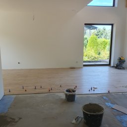 Grzegorz Szczupacki - Układanie płytek podłogowych w jasnym pomieszczeniu z oknem; widoczne poziomice i wiaderka z zaprawą. Częściowo ułożona podłoga, etap prac wykończeniowych.