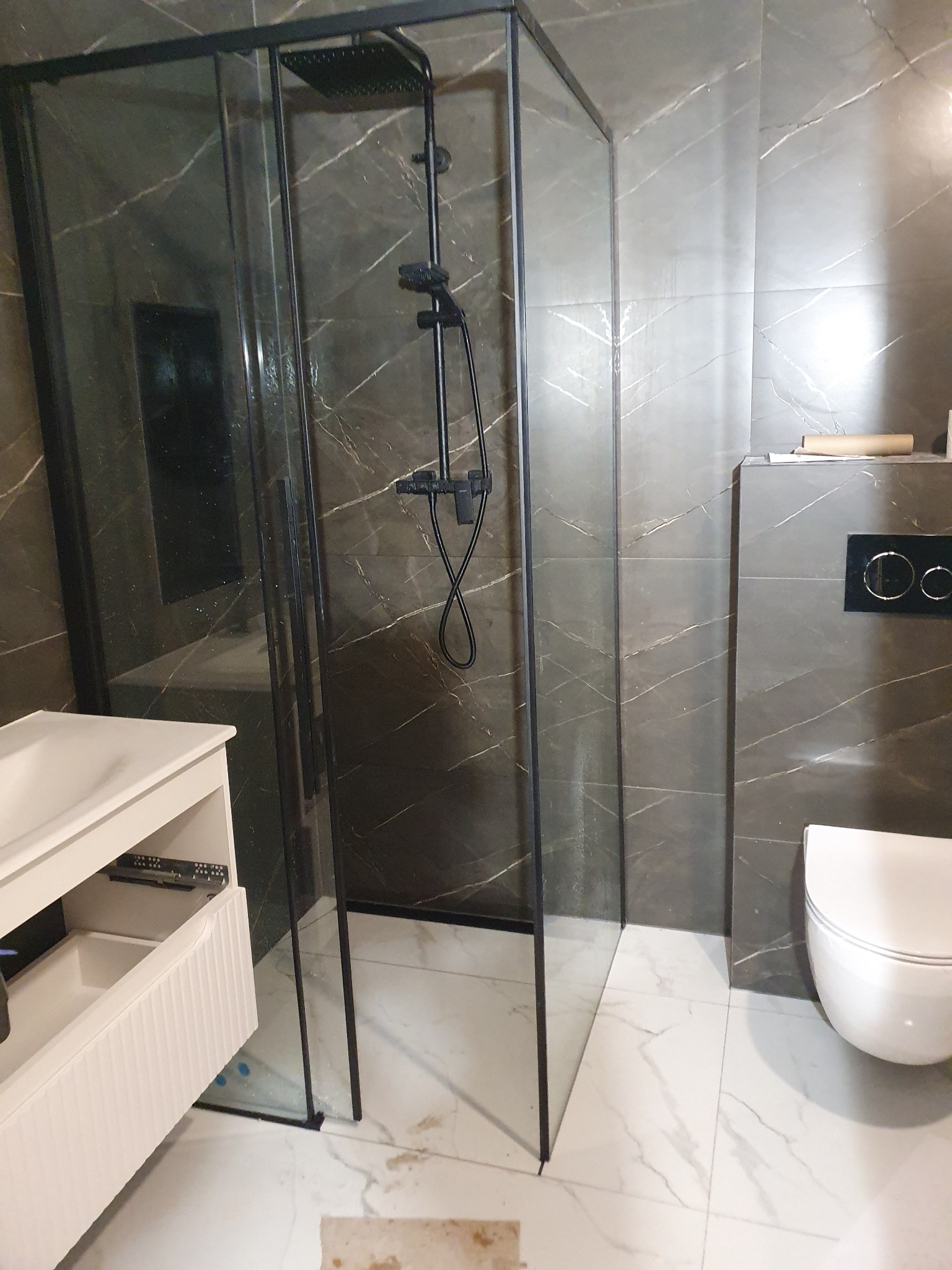 Nowoczesna łazienka z kabiną prysznicową typu walk-in z czarnymi profilami, umywalką z szafką w kolorze złamanej bieli i marmurowymi płytkami na ścianach.