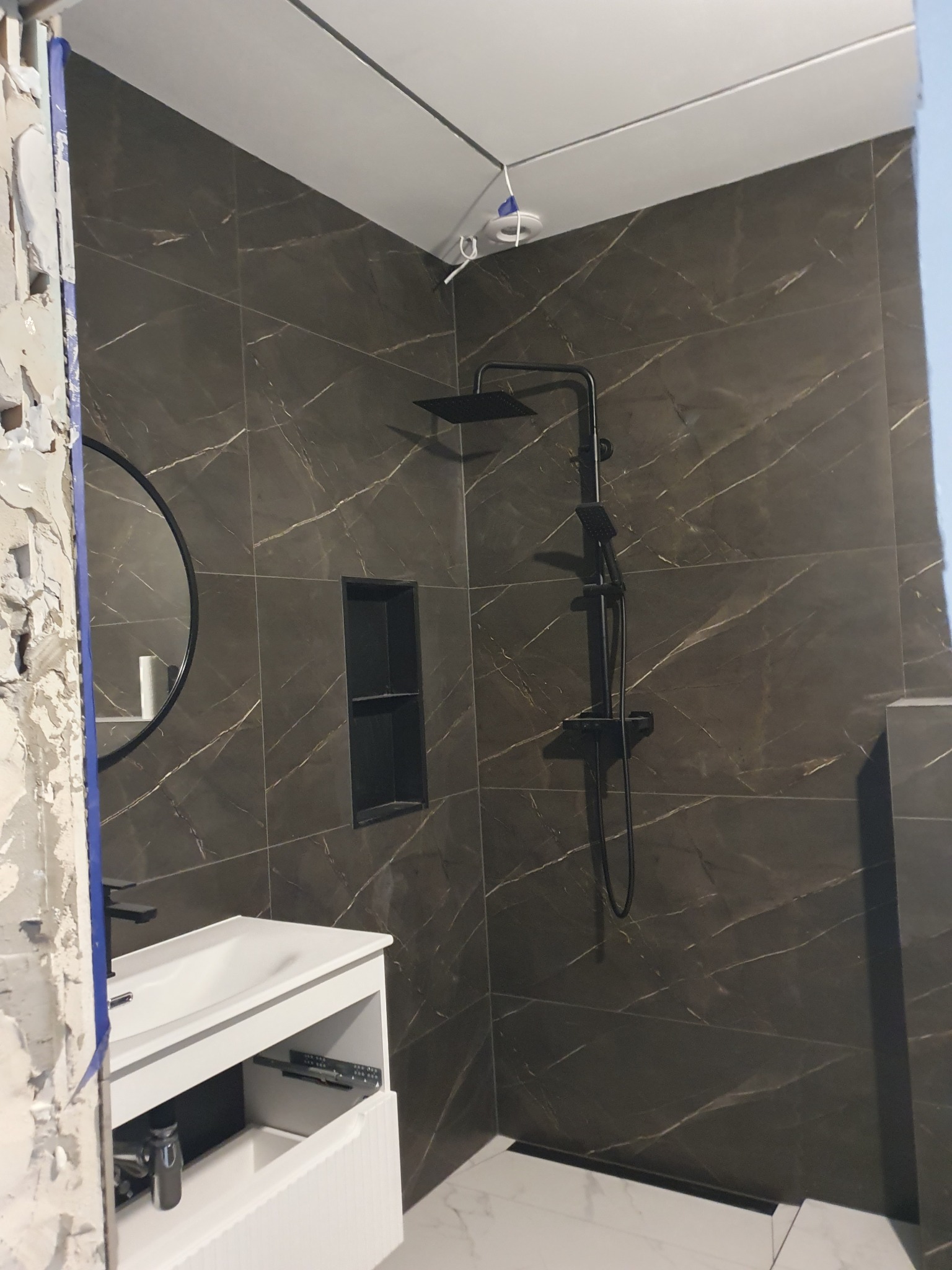 Częściowo wykończona łazienka z ciemnymi, marmurkowymi płytkami, czarną armaturą i białą szafką pod umywalką; widoczne ślady po kuciu na ścianie po lewej stronie.