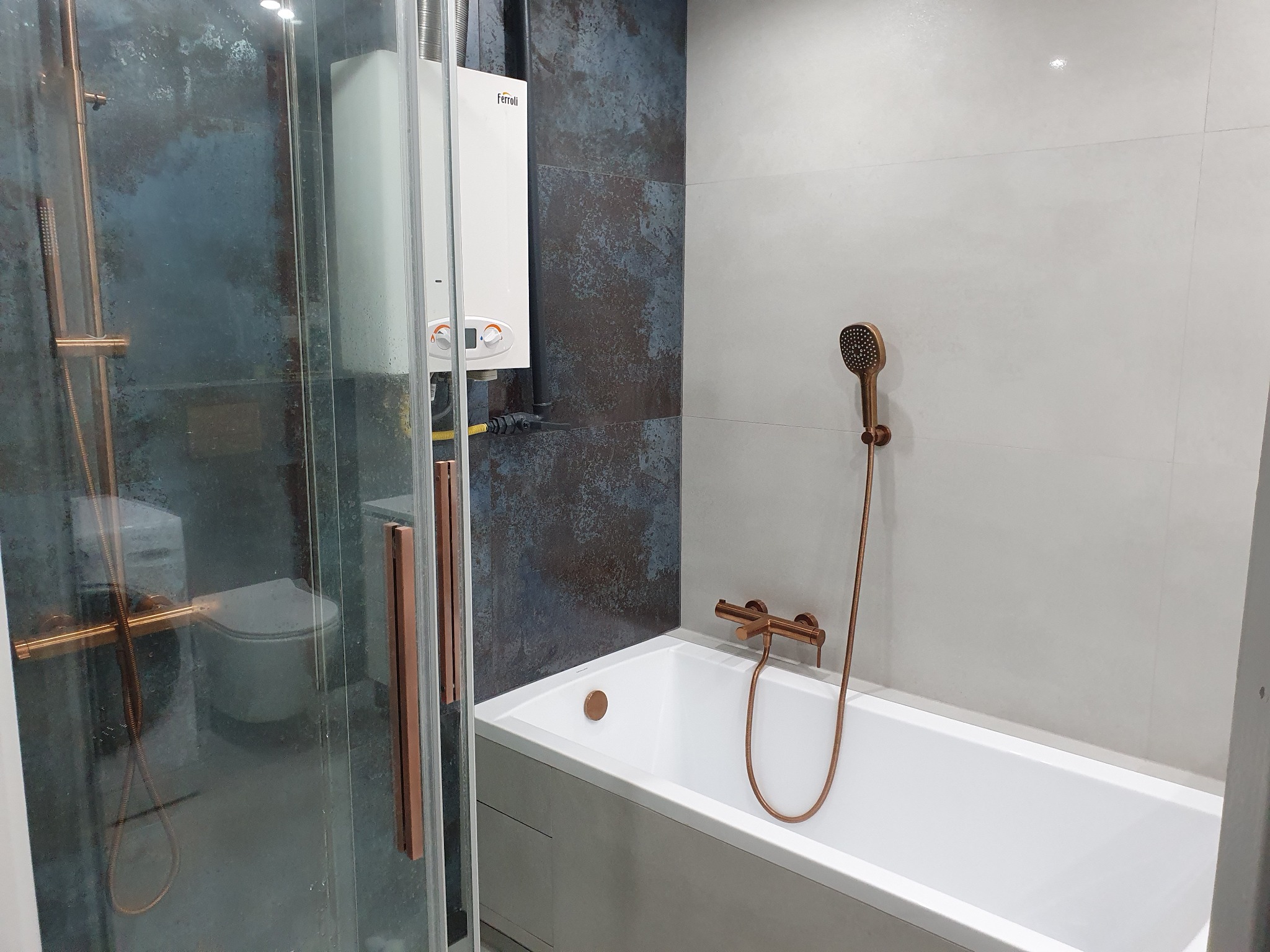 Nowoczesna łazienka z kabiną prysznicową, białą wanną i baterią w kolorze miedzi, na ścianie wiszący piec gazowy Ferroli, płytki w odcieniach szarości i granatu.