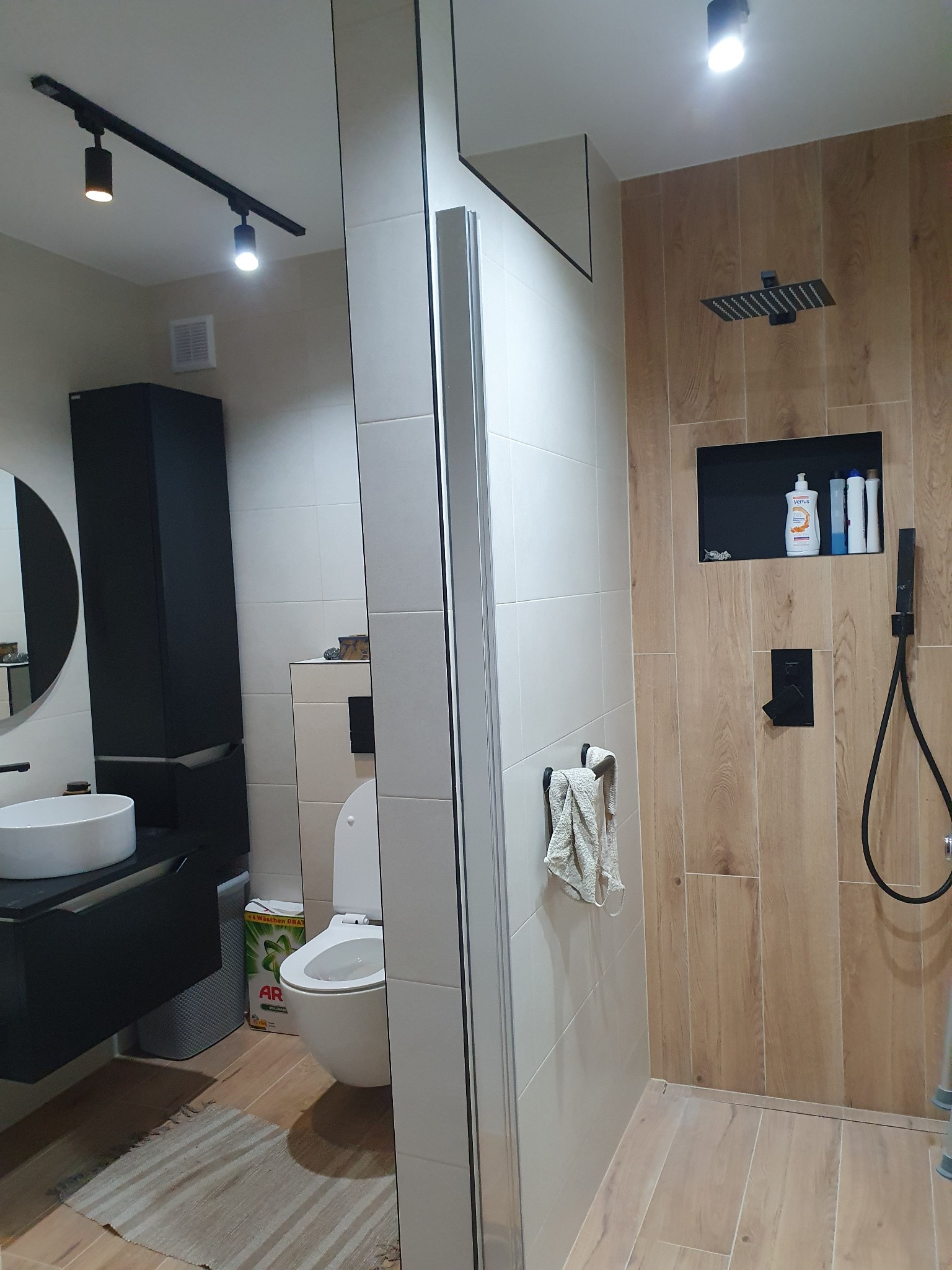 Nowoczesna łazienka z drewnopodobnymi płytkami w strefie prysznica, czarną armaturą, minimalistyczną umywalką nablatową i oświetleniem szynowym.