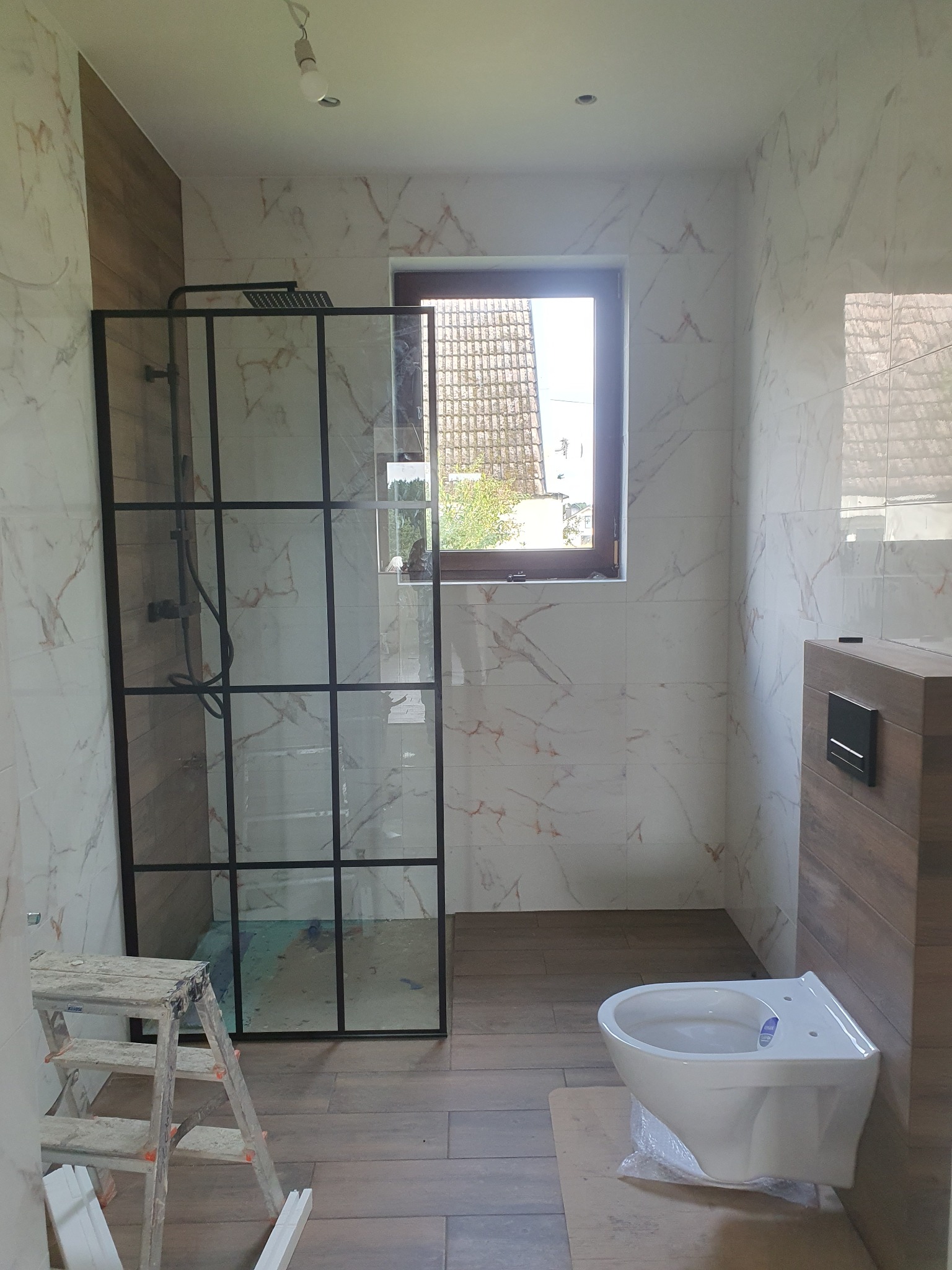 Nowoczesna łazienka z kabiną prysznicową typu walk-in z czarnymi profilami, marmurowymi płytkami na ścianach, drewnopodobną zabudową WC i wiszącą miską ustępową, widoczna drabina malarska.