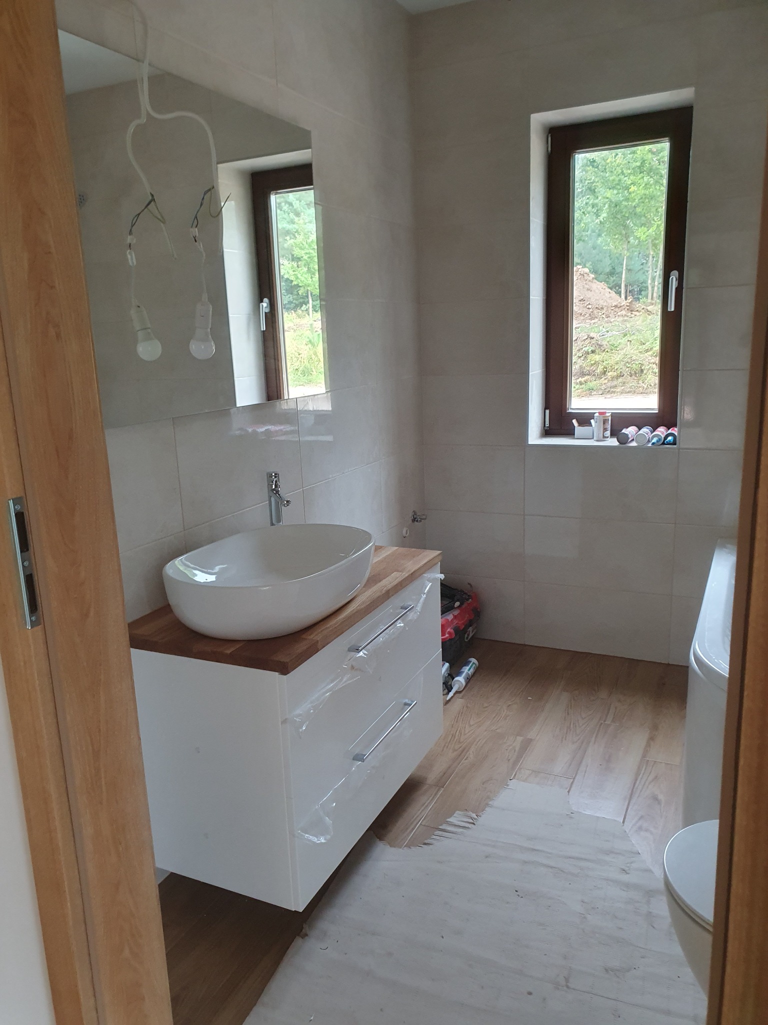 Jasna łazienka w trakcie wykończenia: umywalka nablatowa na szafce, okno z widokiem na zieleń, płytki na ścianach, brakujące oświetlenie, częściowo zakryta folią szafka.