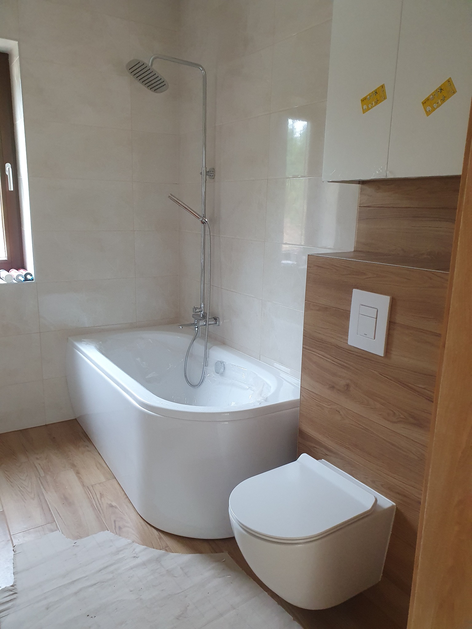 Nowoczesna łazienka z białą wanną, podwieszaną toaletą i panelami imitującymi drewno. Widoczna armatura i płytki w jasnych odcieniach. Częściowo widoczne zabezpieczenia na meblach.