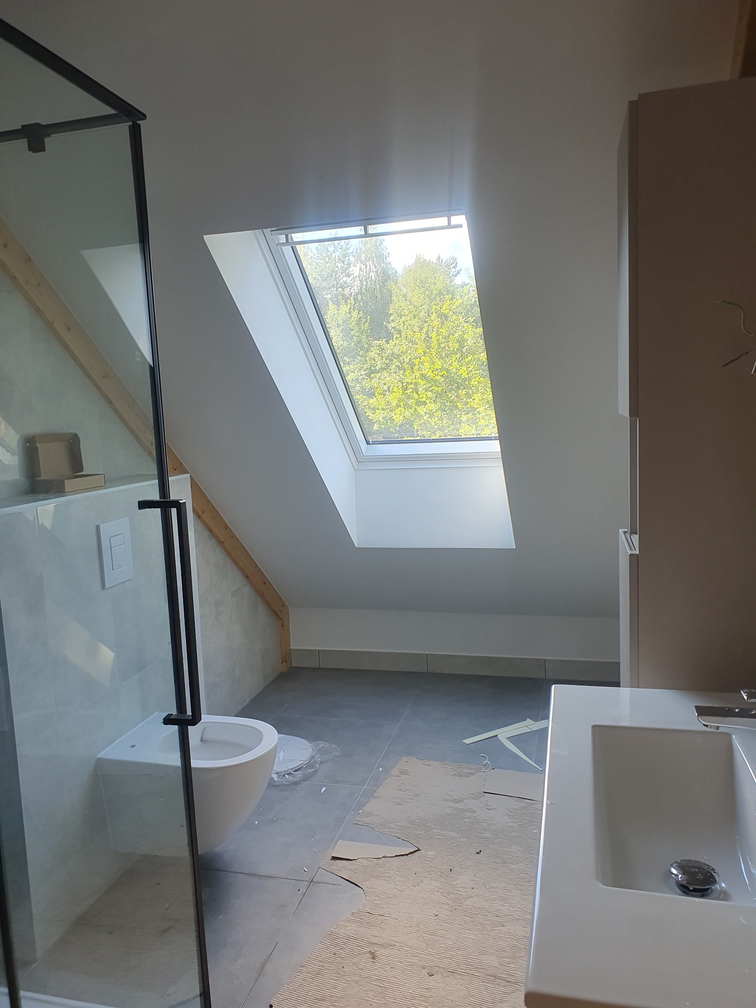 Łazienka na poddaszu ze skośnym oknem dachowym, widok na zieleń. Umywalka, toaleta podwieszana, kabina prysznicowa z czarnymi profilami. Szare płytki podłogowe.
