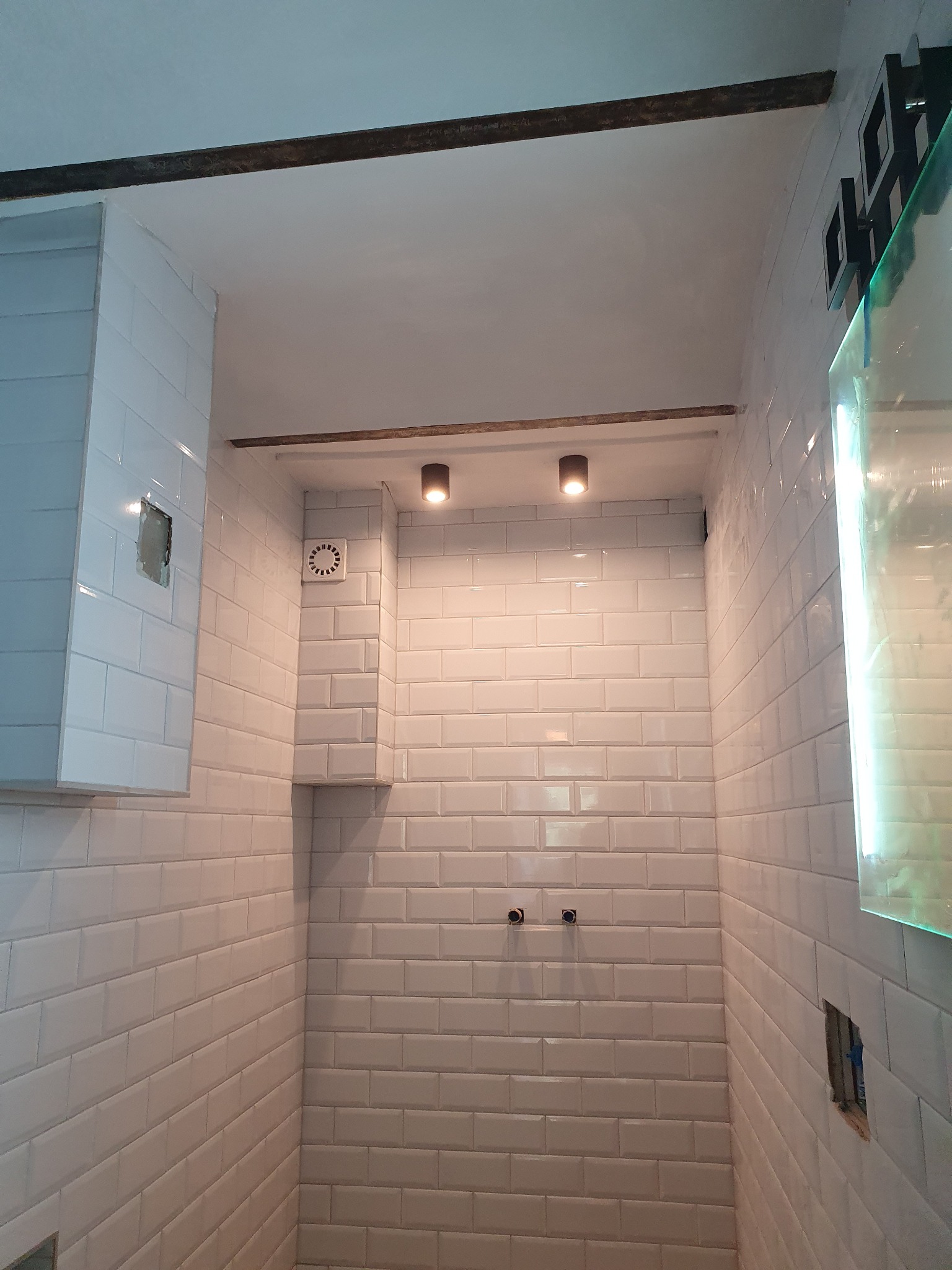 Wykończona łazienka z białymi, prostokątnymi płytkami ułożonymi w cegiełkę, widoczne punkty podłączeń armatury, wentylator i oświetlenie sufitowe.