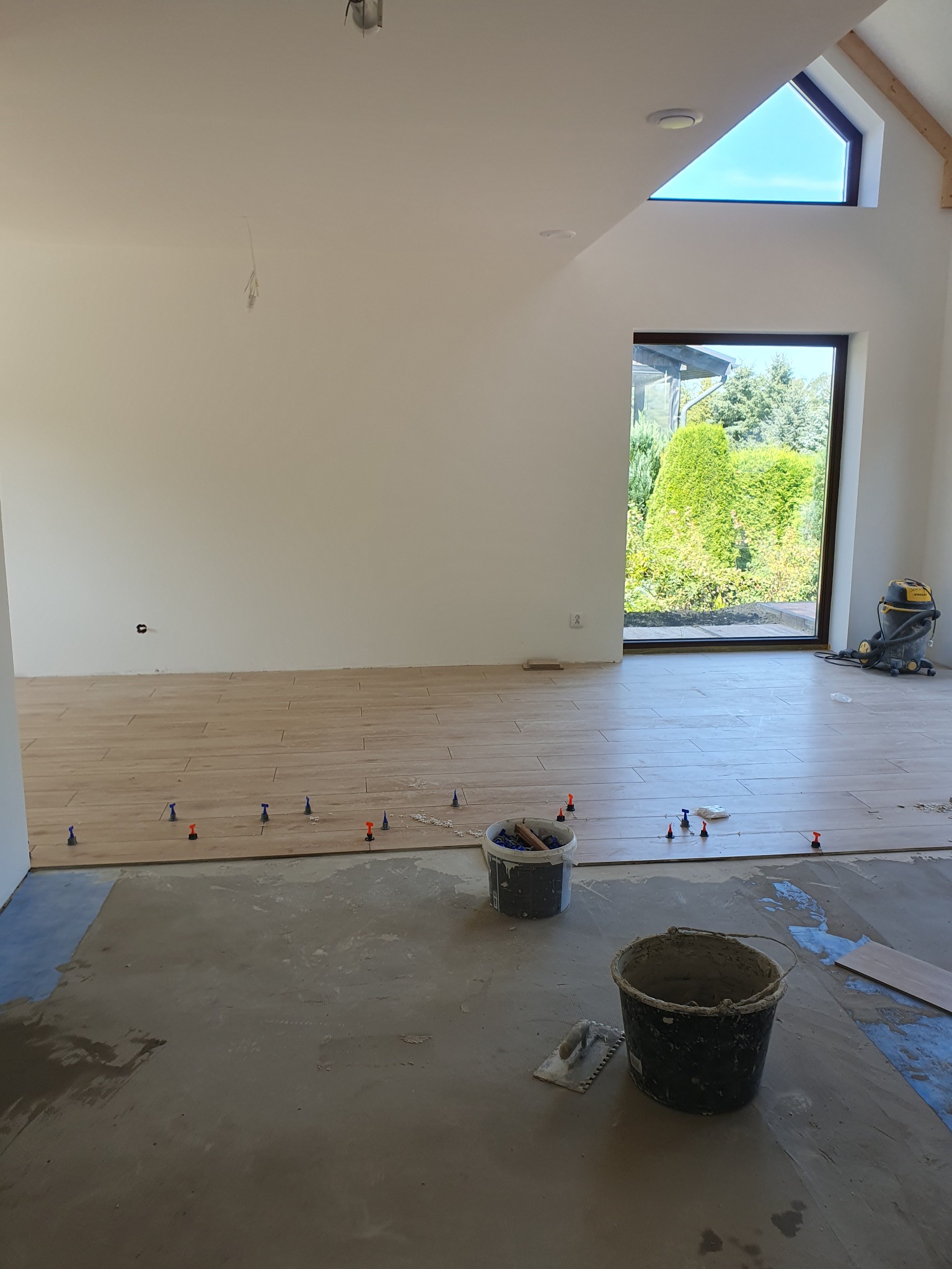 Układanie płytek podłogowych w jasnym pomieszczeniu z oknem; widoczne poziomice i wiaderka z zaprawą. Częściowo ułożona podłoga, etap prac wykończeniowych.
