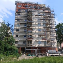 LDV bud PL - Wysoki budynek mieszkalny w trakcie remontu elewacji, otoczony rusztowaniami. Widoczne fragmenty odnowionej fasady i materiały budowlane na dole.