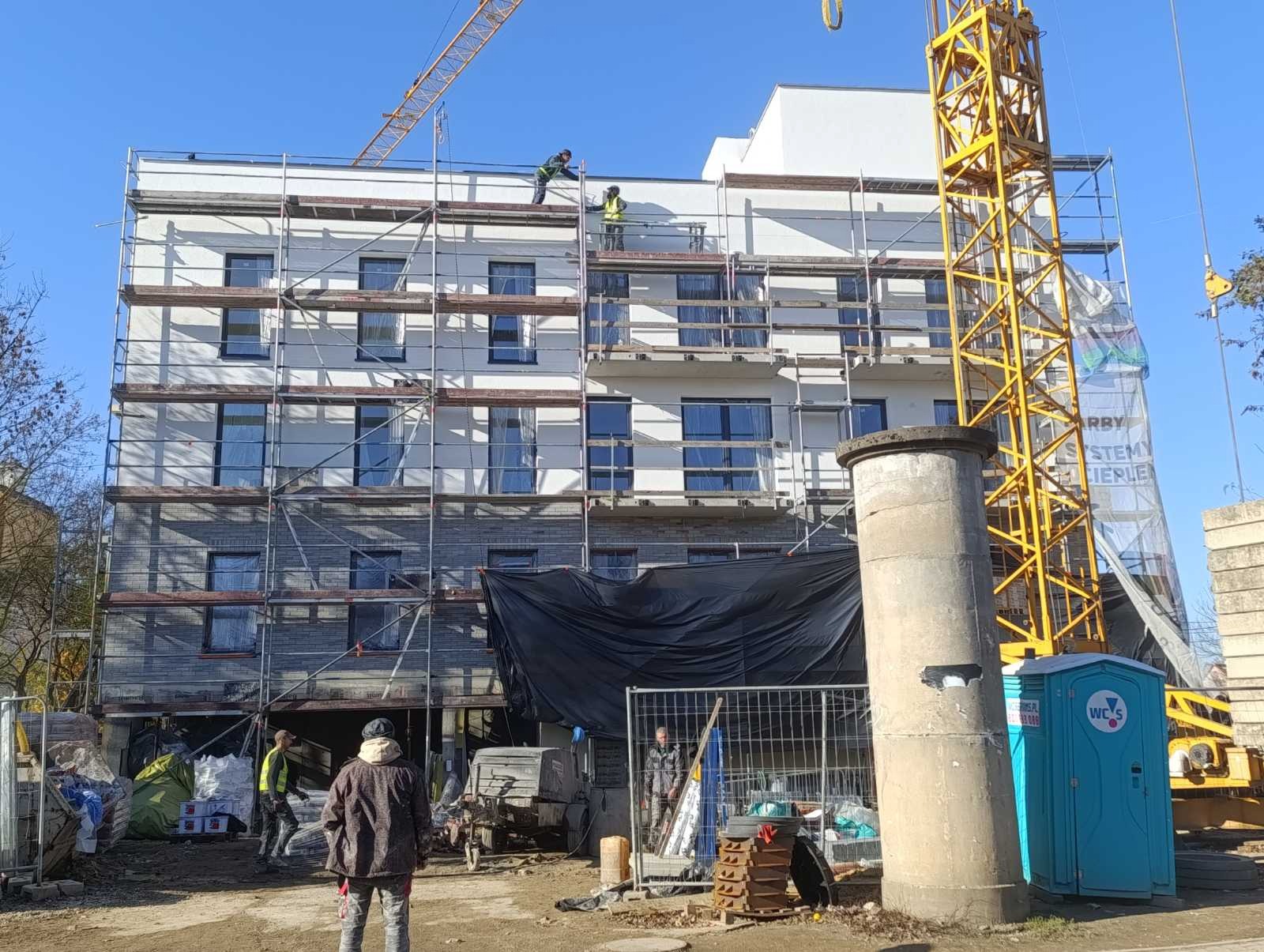 Budynek w trakcie prac wykończeniowych, rusztowania, pracownicy na dachu, żuraw budowlany w tle, widoczny fragment terenu budowy z przenośną toaletą.