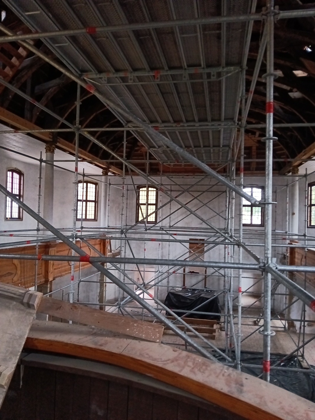 Wnętrze remontowanego budynku z drewnianym stropem, widoczne rusztowania i okna. Prace przygotowawcze do malowania ścian w zabytkowym obiekcie.