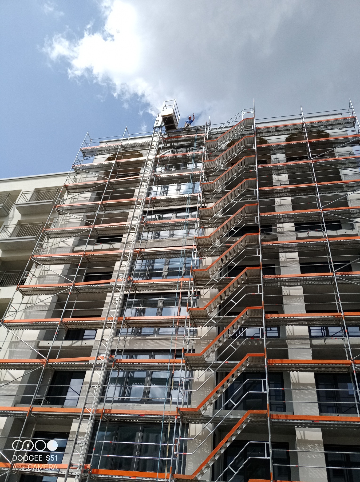 Budynek w trakcie remontu, osłonięty rusztowaniami z pomarańczowymi elementami. Widoczni pracownicy na górze, błękitne niebo z chmurami w tle. Ujęcie z dołu.