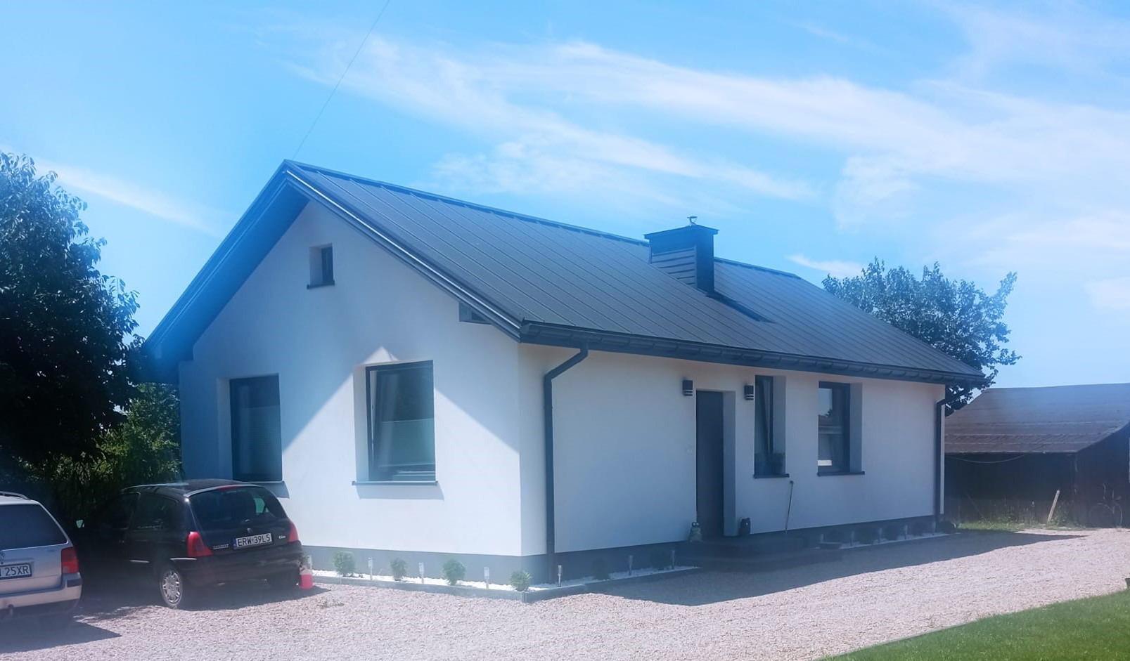 Nowoczesny, parterowy dom z ciemnym dachem i jasną elewacją, otoczony żwirowym podjazdem i zielenią, widziany w słoneczny dzień.
