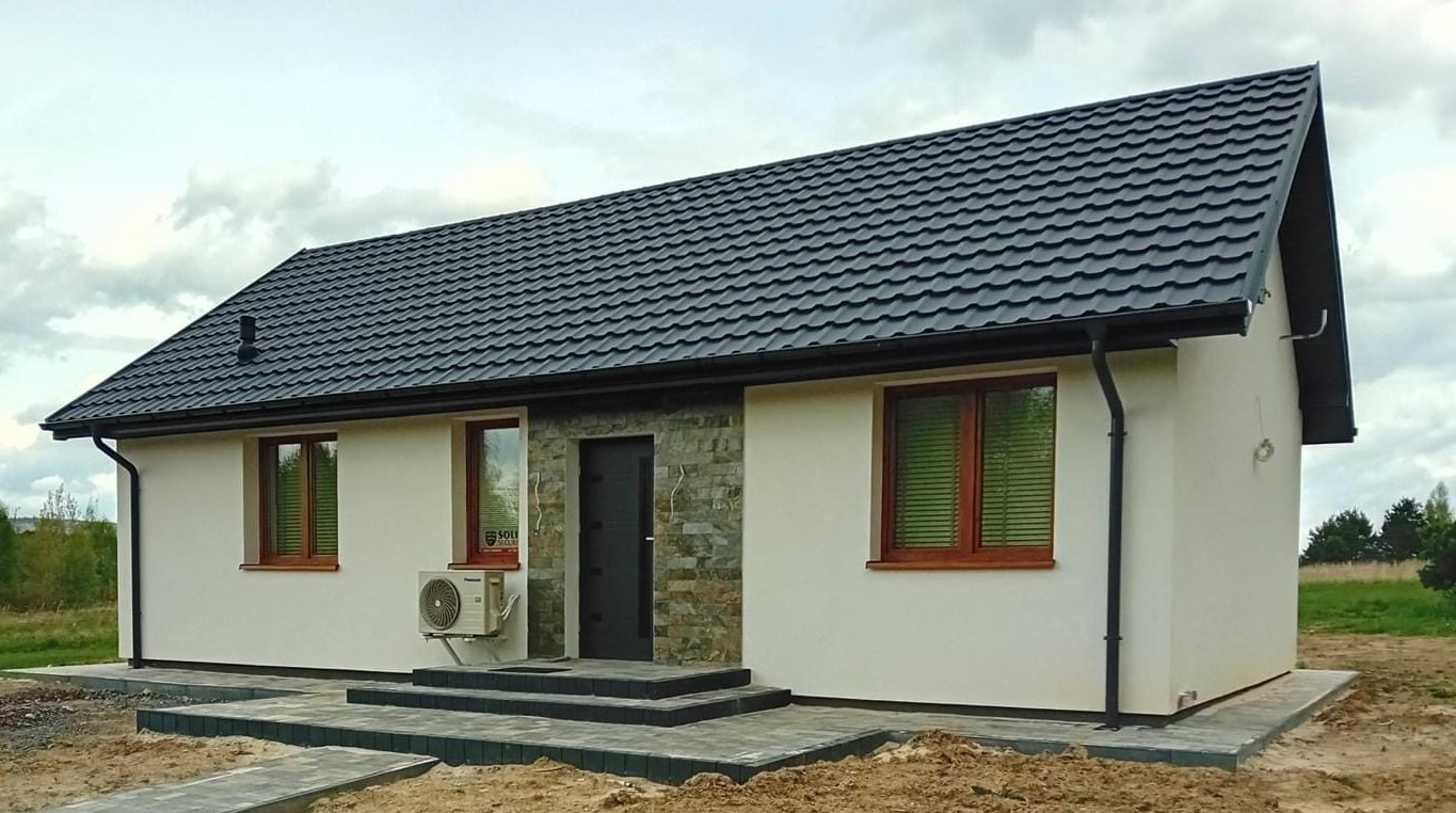 Gotowy dom szkieletowy z czarną dachówką, jasną elewacją, kamiennym wykończeniem przy wejściu i zamontowaną klimatyzacją na bocznej ścianie.