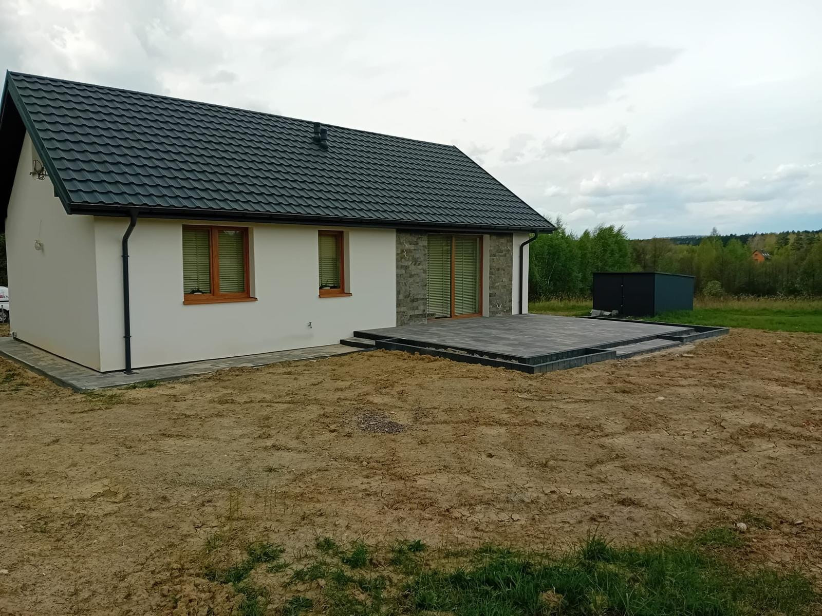 Nowoczesny dom szkieletowy z ciemnym dachem i tarasem z szarej kostki, widok z zewnątrz na tle pochmurnego nieba i zieleni drzew.