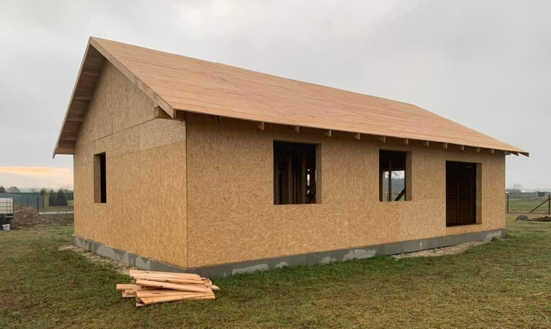 Surowy stan domu z płyt OSB z widocznymi otworami okiennymi i drzwiowymi, na trawiastym terenie, z deskami ułożonymi obok.