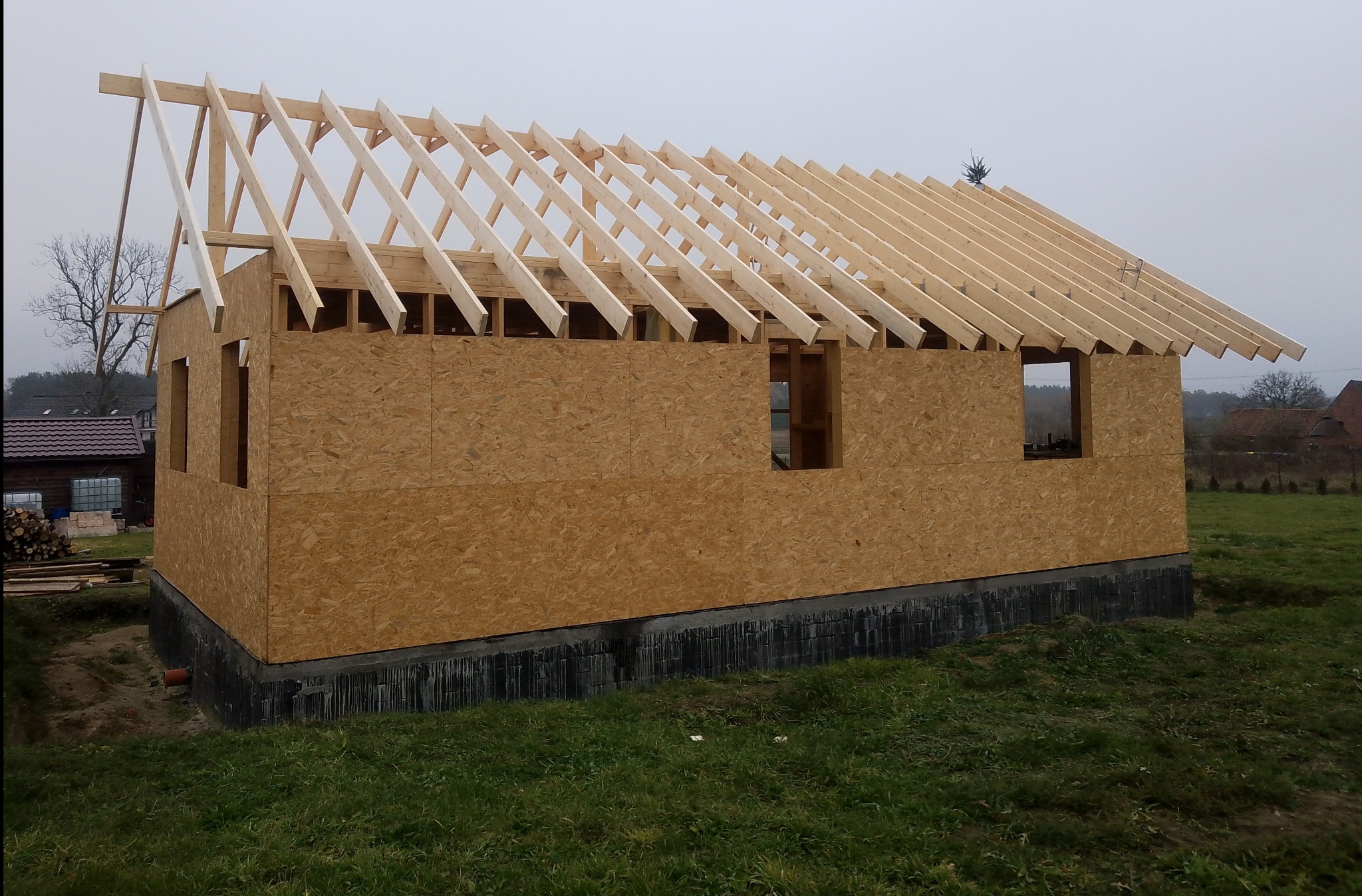 Surowy stan budowy domu szkieletowego z widocznymi ścianami z płyt OSB, drewnianą konstrukcją dachu oraz fundamentami, na tle pochmurnego nieba i zielonej trawy.