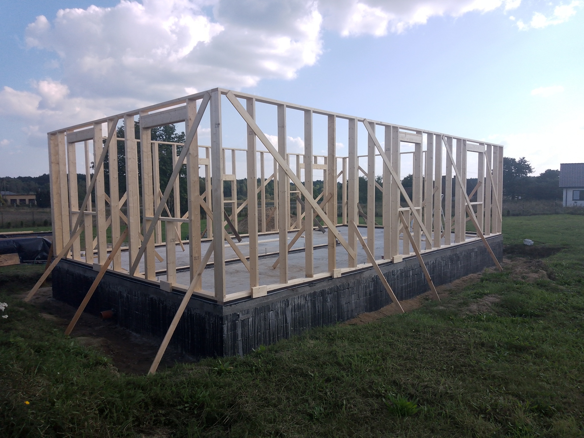 Surowy szkielet konstrukcji drewnianej w Tychowie, oparty na betonowym fundamencie, z widocznymi stężeniami i podporami, na tle zielonej trawy i błękitnego nieba z chmurami.