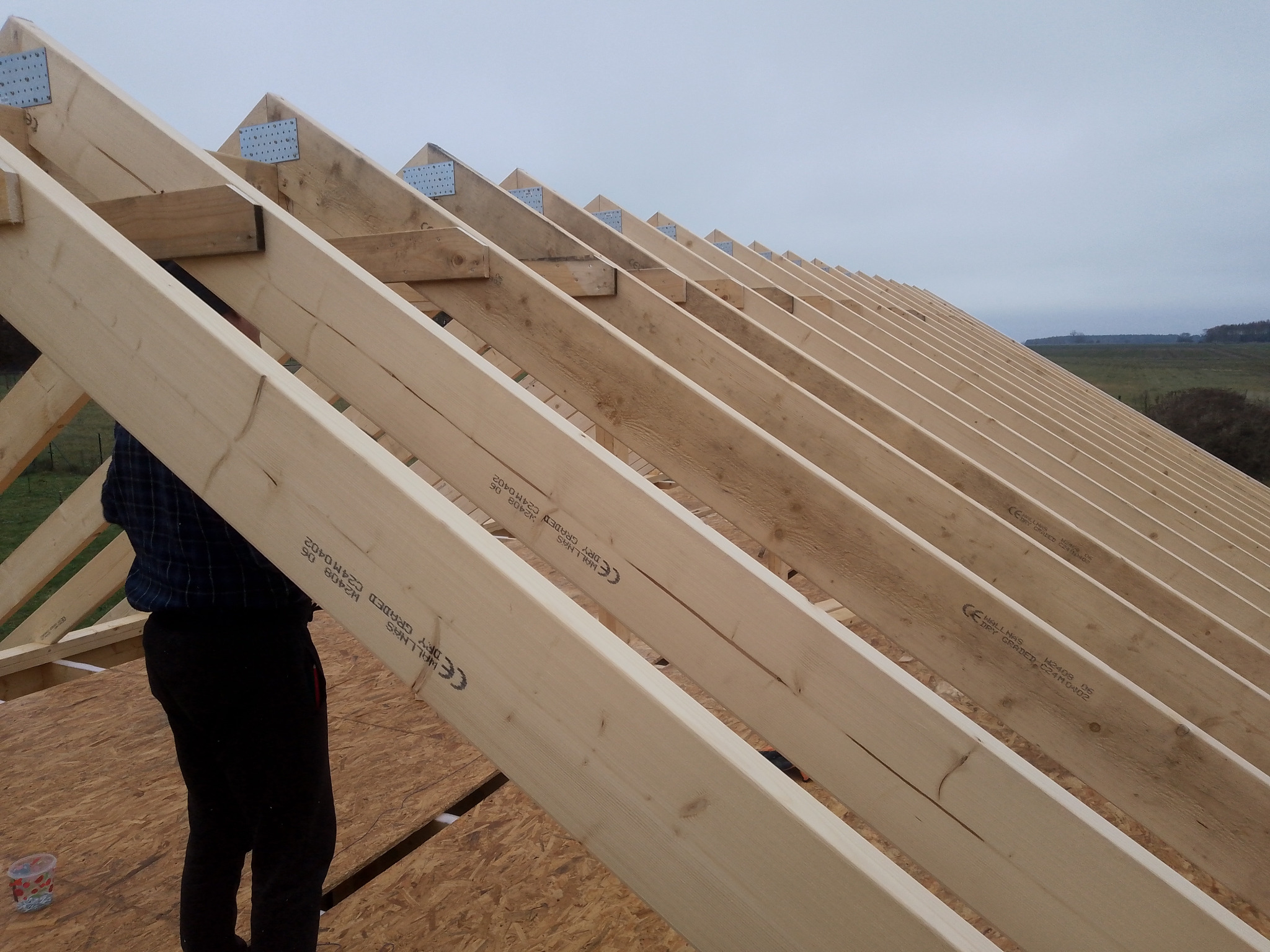 Konstrukcja drewniana dachu w trakcie budowy, widoczne krokwie i łączniki, w tle krajobraz, częściowo widoczna sylwetka osoby w ciemnych spodniach i kraciastej koszuli.