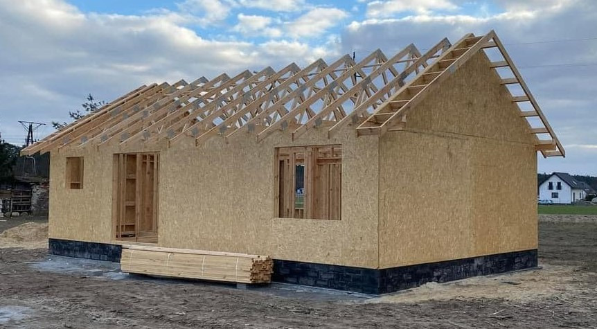 Surowy stan domu w konstrukcji szkieletowej z widocznym poszyciem OSB ścian zewnętrznych i konstrukcją więźby dachowej, stos drewna na ziemi, w tle zabudowa jednorodzinna.