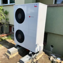 Biała pompa ciepła Saunier Duval GeniaAir Split zamontowana na betonowych fundamentach przy zielonym budynku z widocznymi oknami.