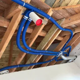 Niebieskie rury wentylacyjne przymocowane do drewnianej konstrukcji stropu, widoczne elementy montażowe i fragment sufitu.