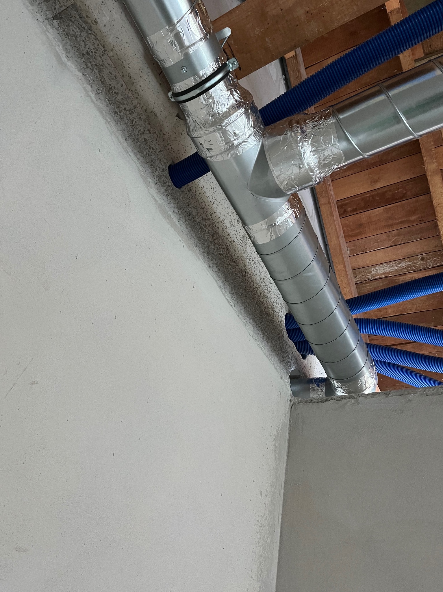 Fragment instalacji wentylacyjnej z rurami spiro, zaizolowanymi taśmą aluminiową, oraz niebieskimi przewodami elastycznymi, zamontowanej pod drewnianym stropem w budynku.
