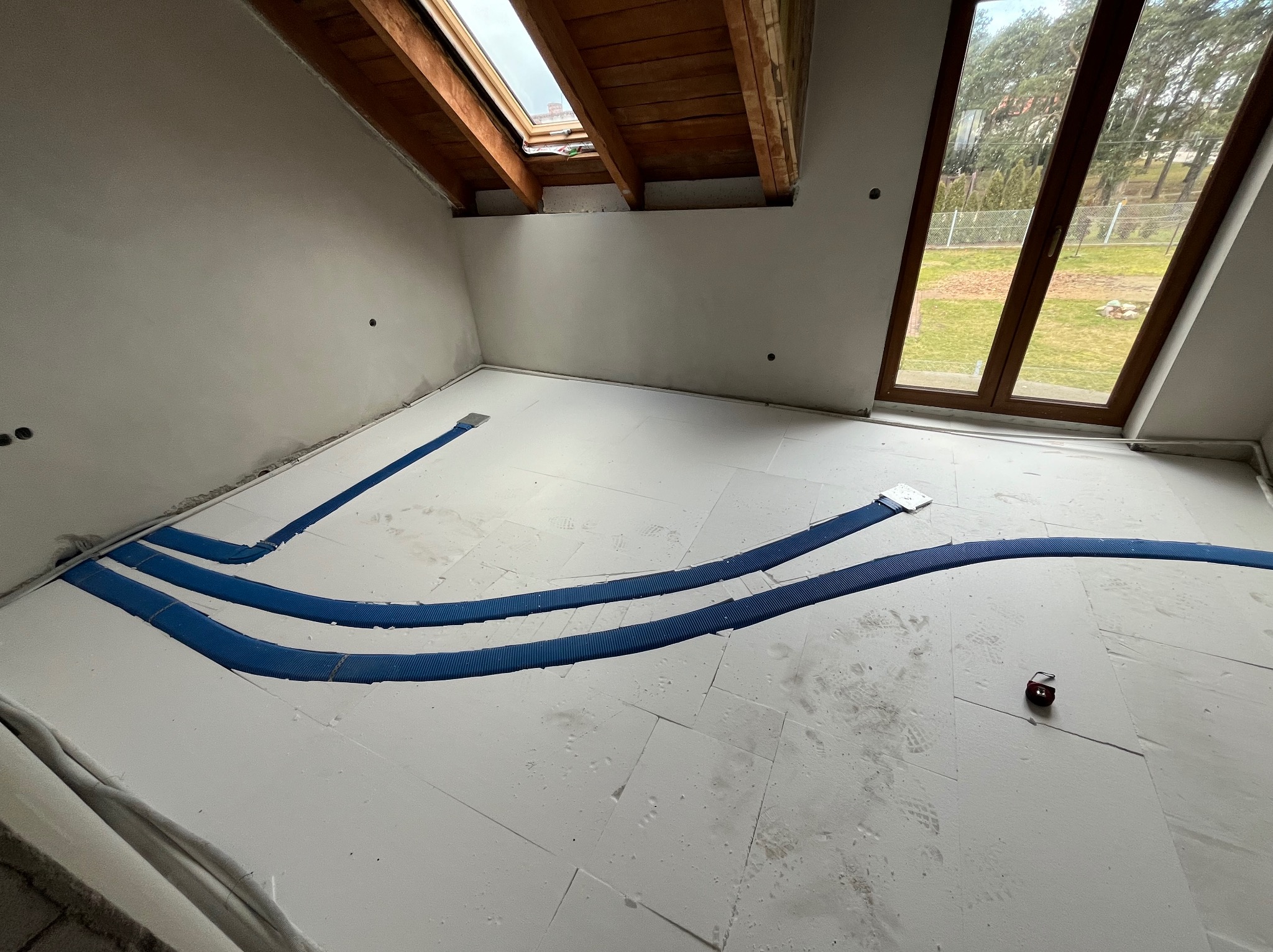 Styropianowe płyty izolacyjne podłogi na poddaszu z ułożonymi niebieskimi rurami systemu rozprowadzania powietrza, widoczne okno dachowe i okno na ścianie szczytowej.