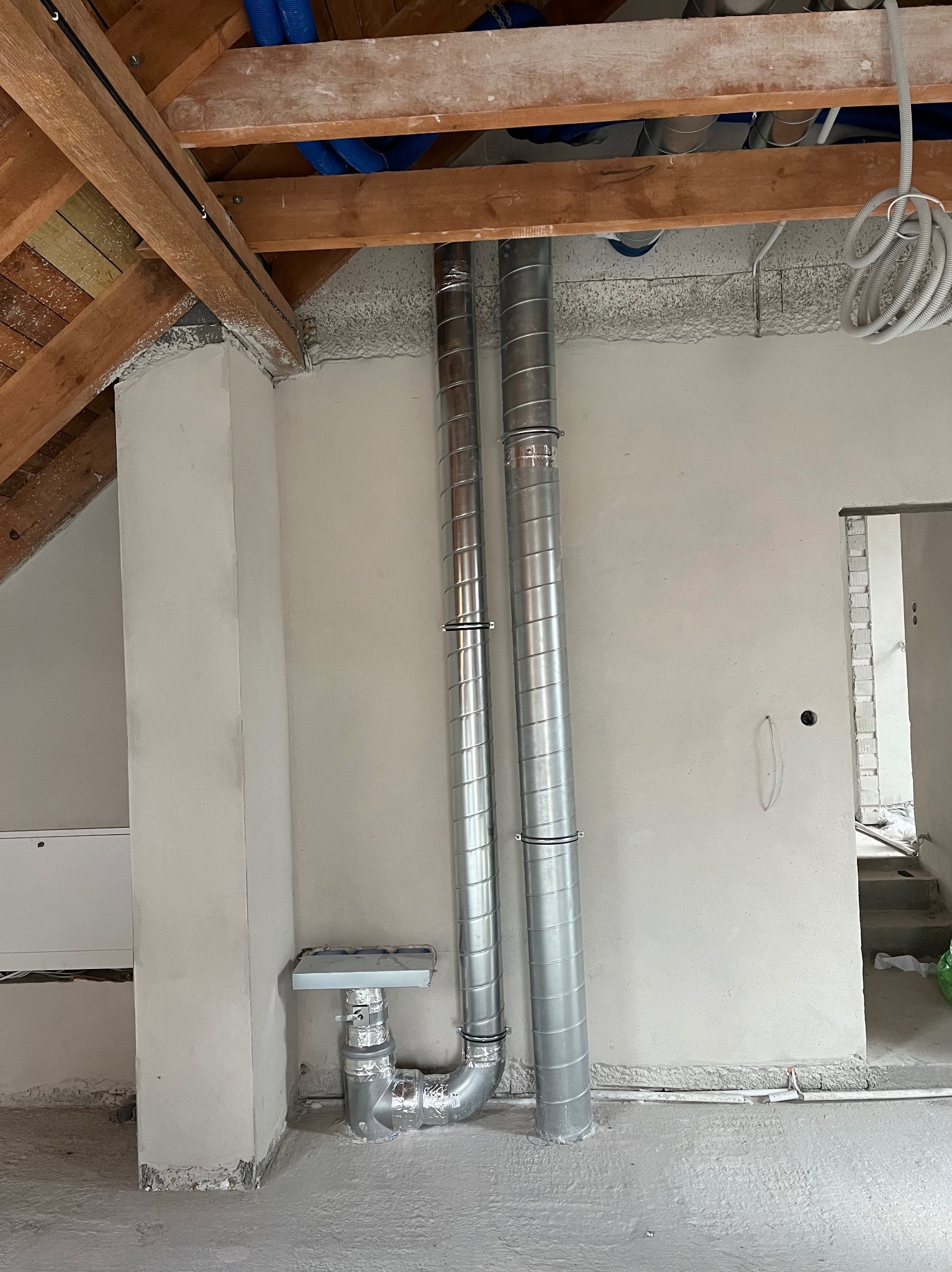 Dwie pionowe, metalowe rury wentylacyjne z widocznymi łączeniami, prowadzone w surowym wnętrzu poddasza z drewnianą konstrukcją stropu i nieotynkowanymi ścianami, instalacja w trakcie montażu.