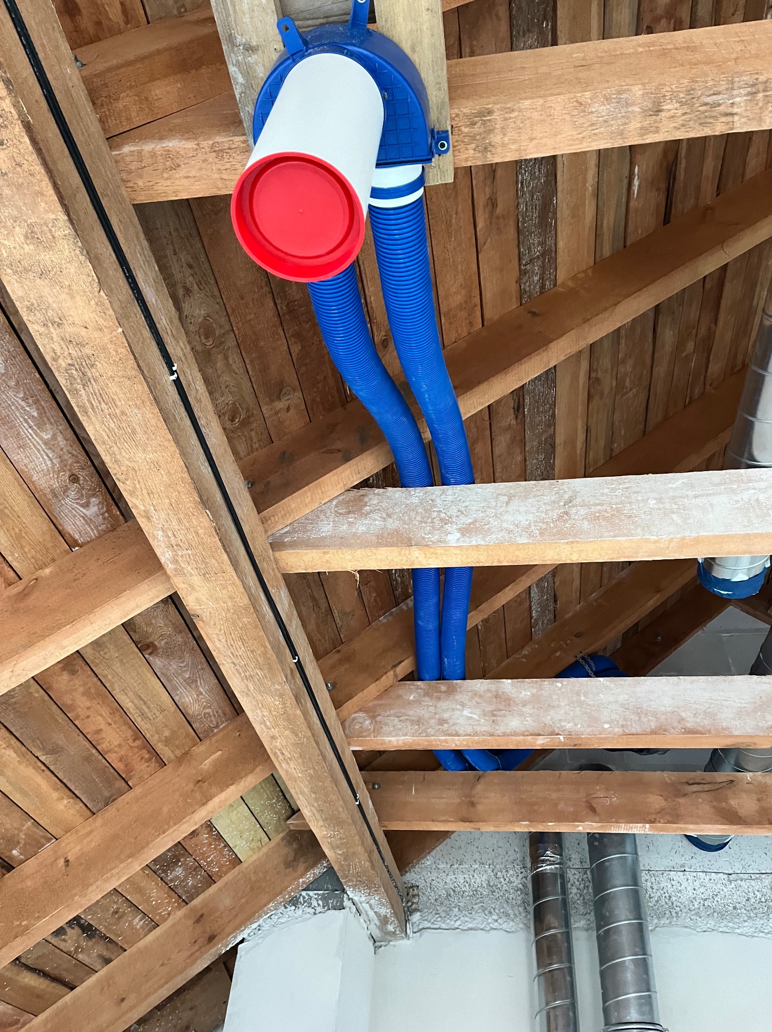 Instalacja systemu wentylacji z widocznymi niebieskimi przewodami w przestrzeni poddasza z drewnianą konstrukcją stropu i fragmentami instalacji metalowej.