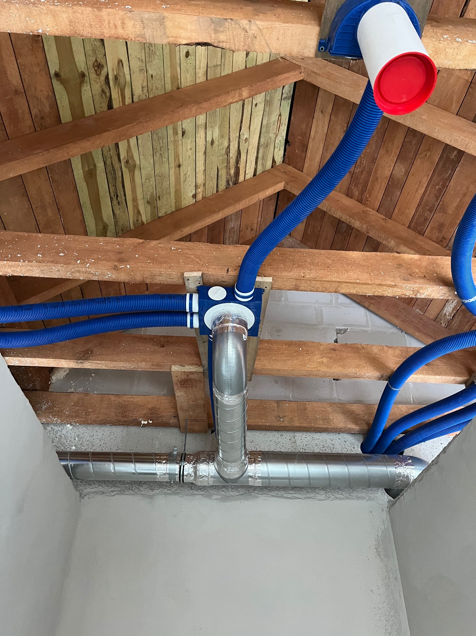 Widok od dołu na instalację wentylacyjną z niebieskimi rurami karbowanymi i srebrnymi kanałami w surowym wnętrzu poddasza z drewnianą konstrukcją stropu.