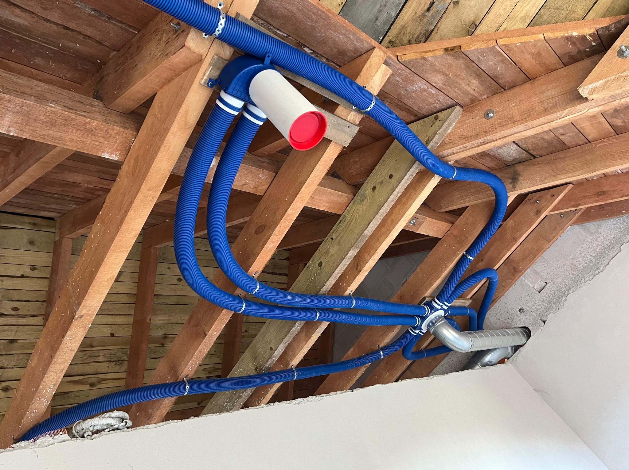 Niebieskie rury wentylacyjne przymocowane do drewnianej konstrukcji stropu, widoczne elementy montażowe i fragment sufitu.