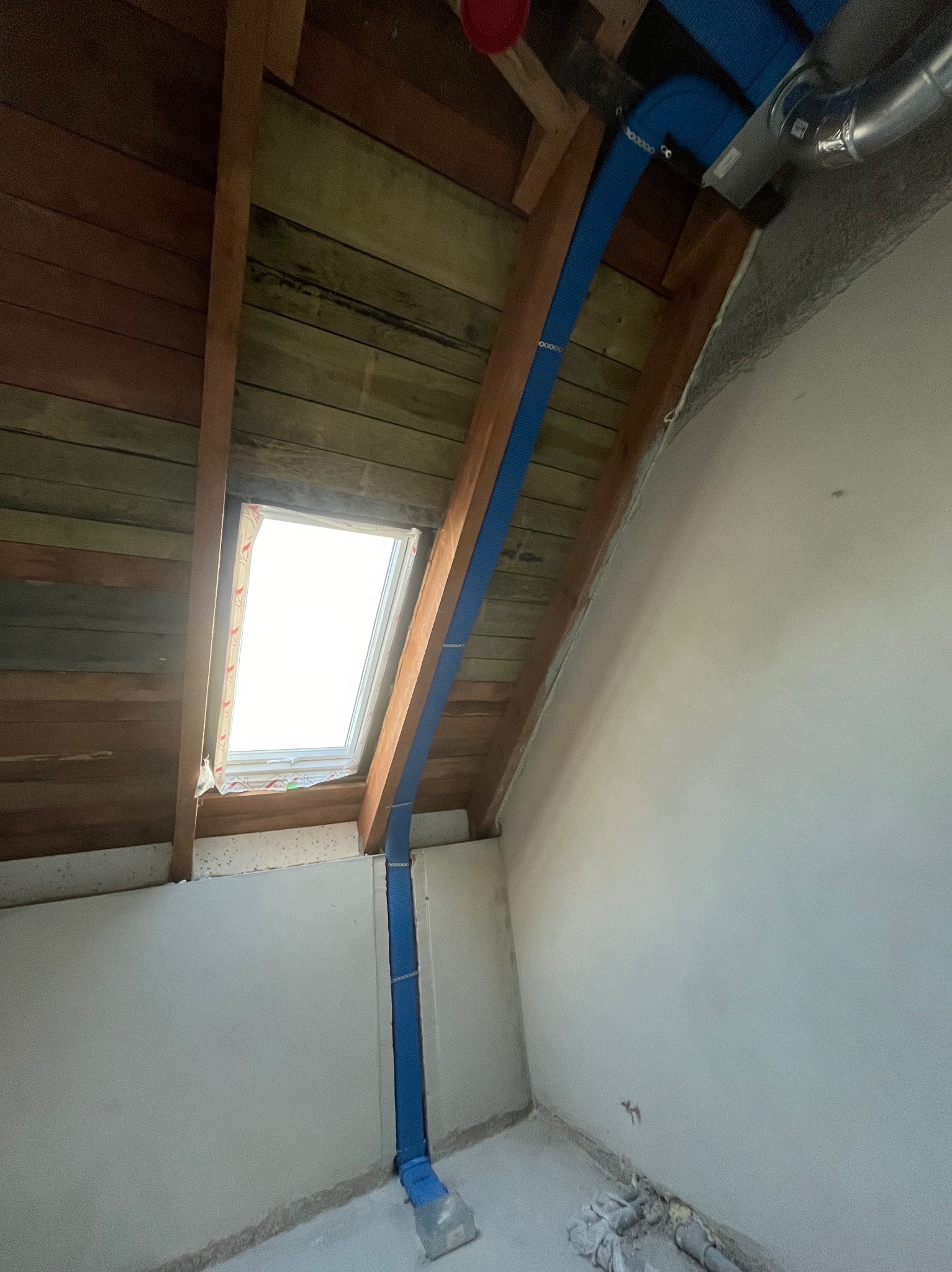 Niebieski przewód rekuperacji biegnący od sufitowego okna do kanału wentylacyjnego w poddaszu z drewnianą konstrukcją i oknem dachowym.