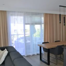 Beżowe zasłony zaciemniające i białe firany w przestronnym salonie z wyjściem na balkon, widoczne z bliska z boku szarej sofy i drewnianego stołu z czarnymi nogami.