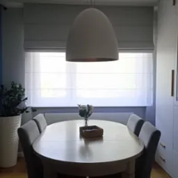 Jasne wnętrze jadalni z owalnym stołem, szarymi krzesłami i roletą rzymską w oknie, oświetlone wiszącą lampą w kształcie kopuły.