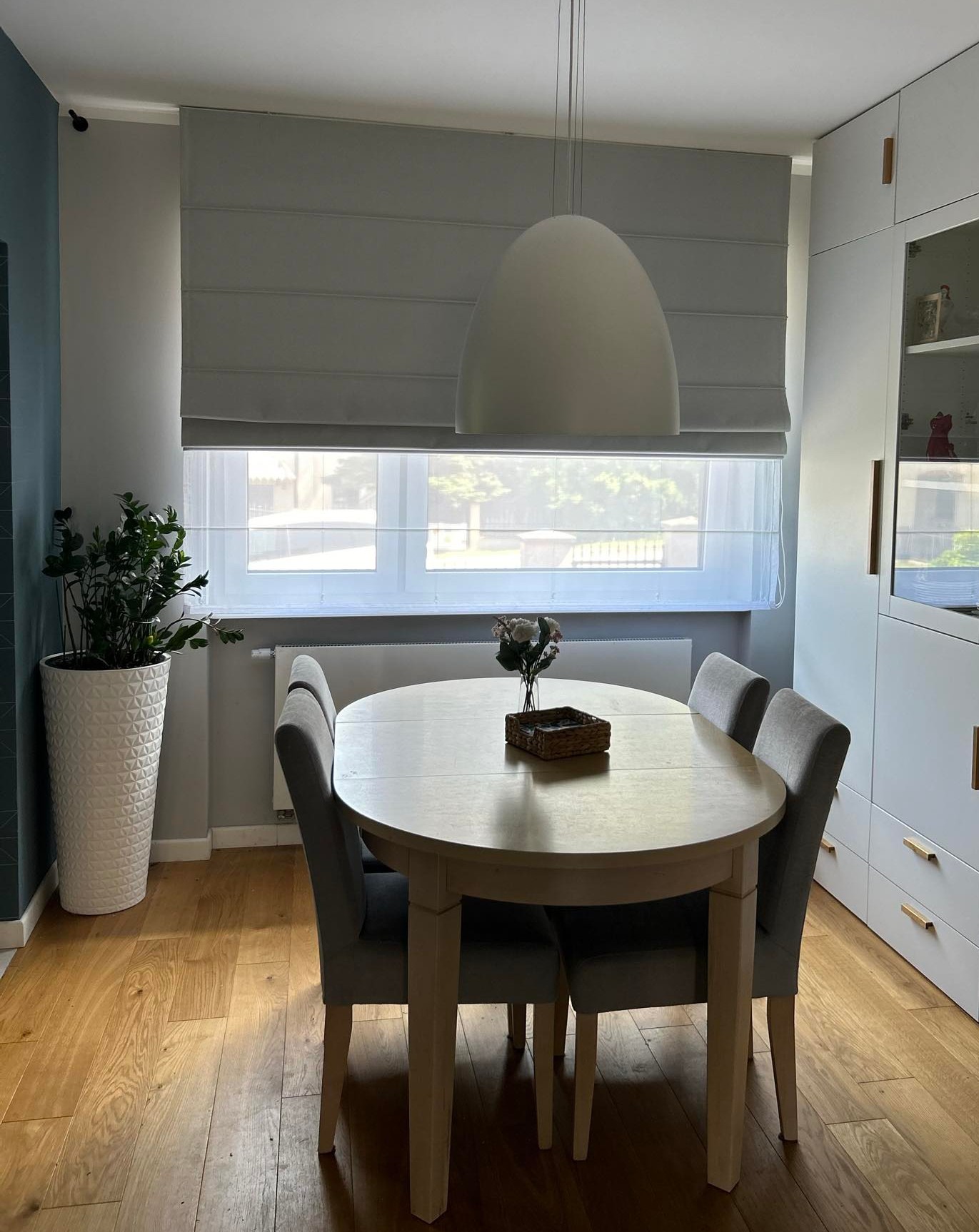 Jasnoszara roleta rzymska zamontowana w oknie nad okrągłym stołem jadalnym z szarymi krzesłami, oświetlonym przez wiszącą lampę w kształcie kopuły.