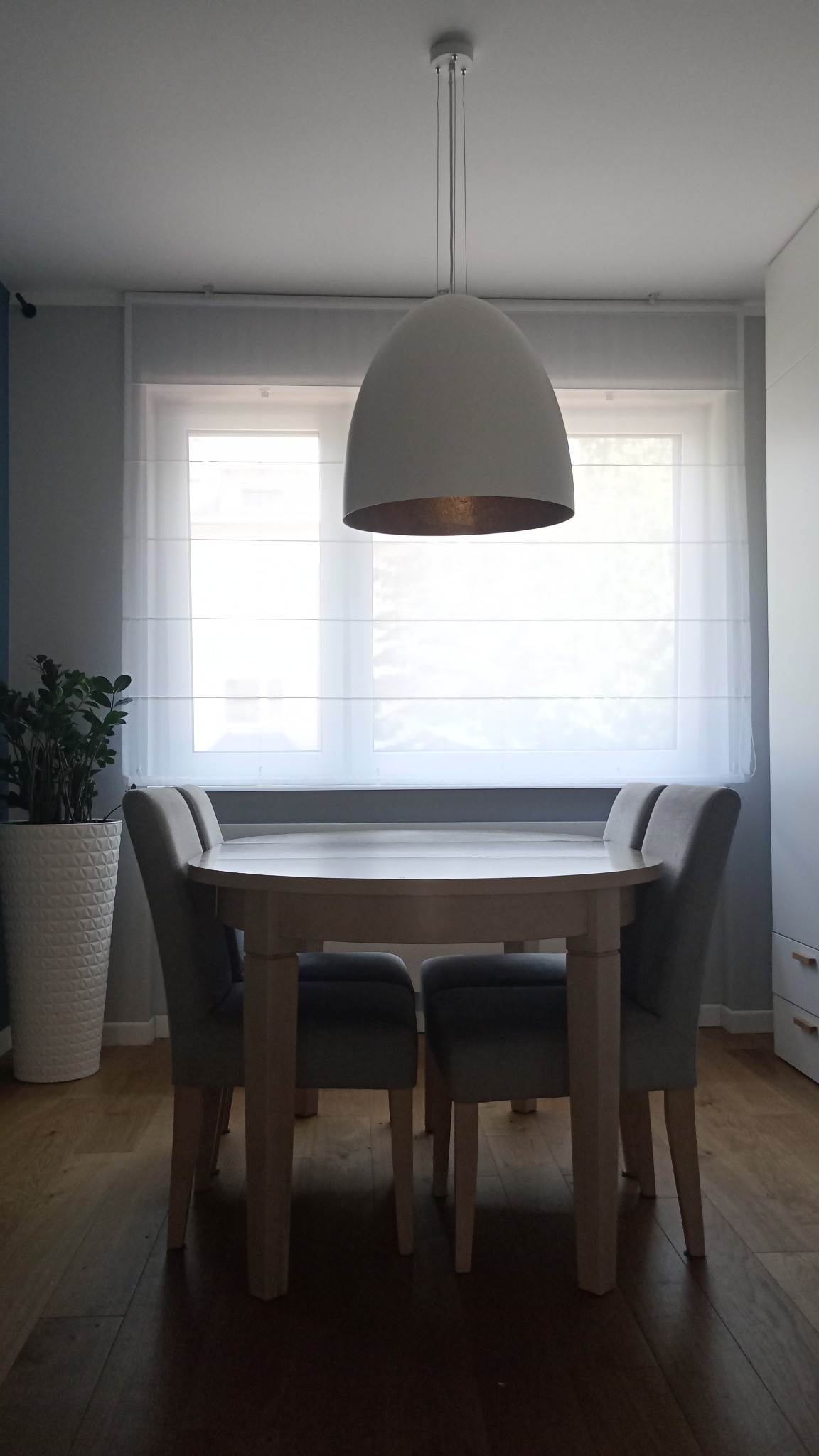 Jasna jadalnia z okrągłym stołem, krzesłami i roletą rzymską w oknie, oświetlona nowoczesną lampą wiszącą.