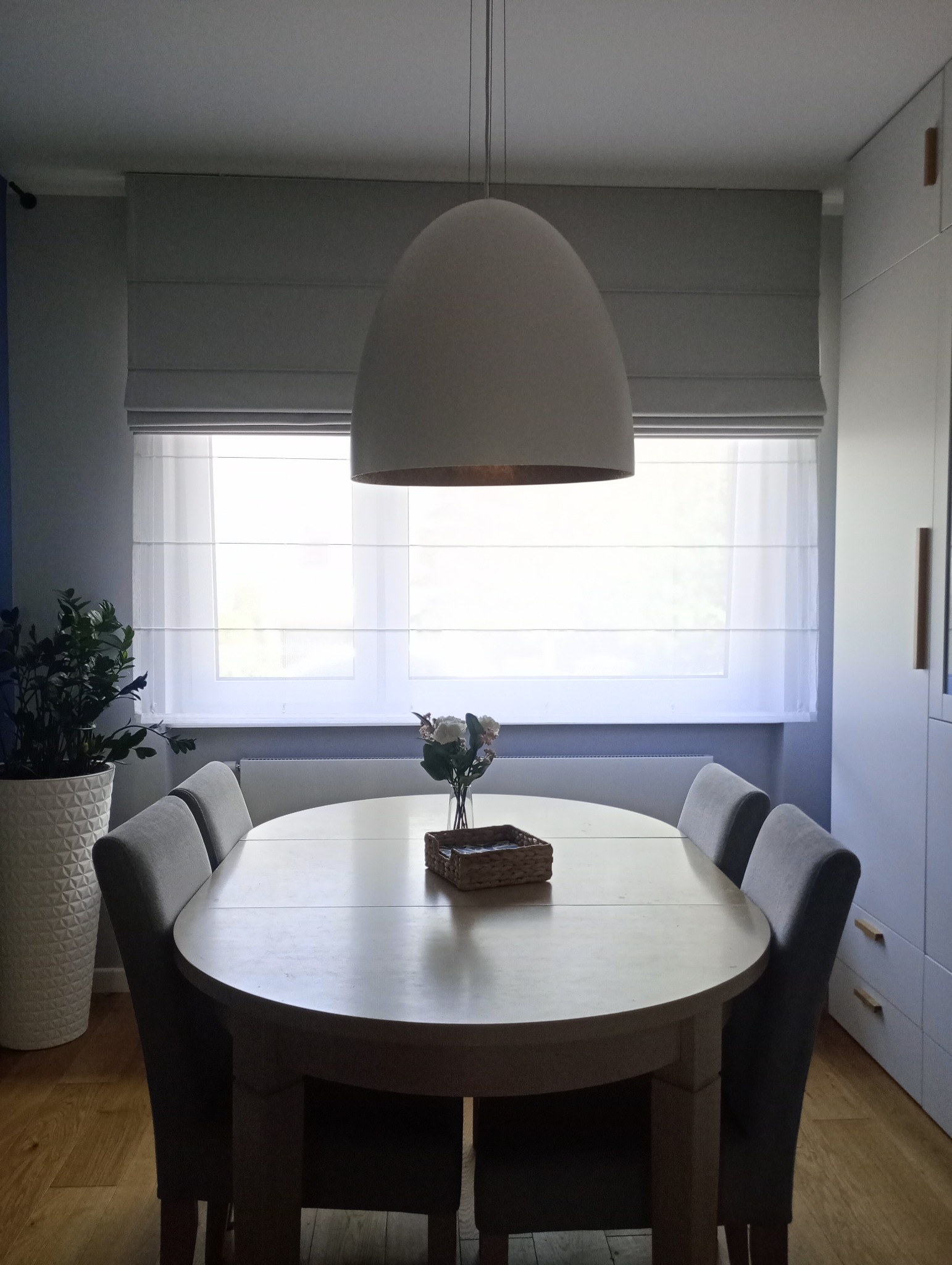 Jasne wnętrze jadalni z owalnym stołem, szarymi krzesłami i roletą rzymską w oknie, oświetlone wiszącą lampą w kształcie kopuły.