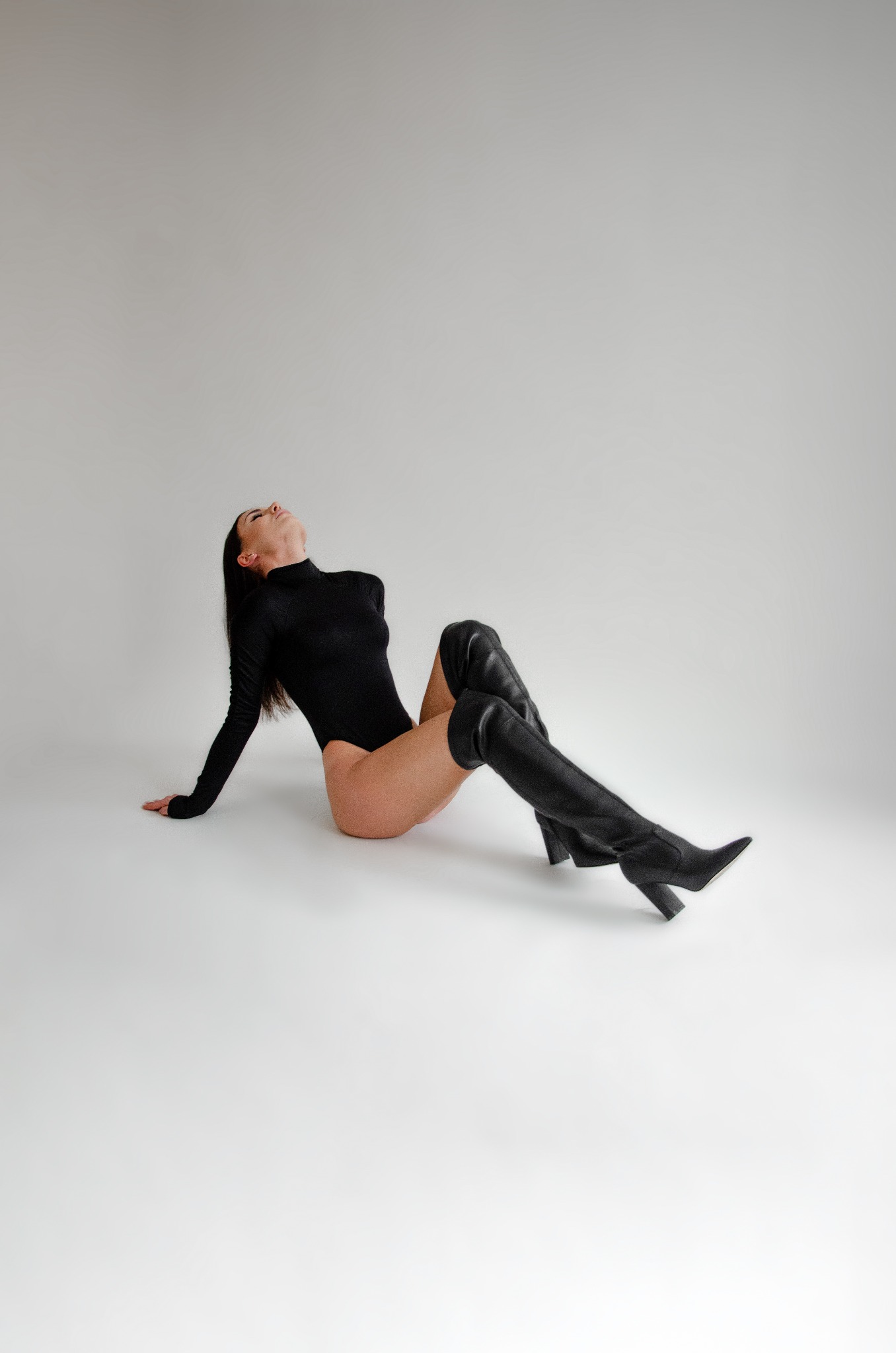 Kobieta w czarnym body i wysokich skórzanych kozakach na obcasie, pozująca na jasnym tle, z głową odchyloną do tyłu.