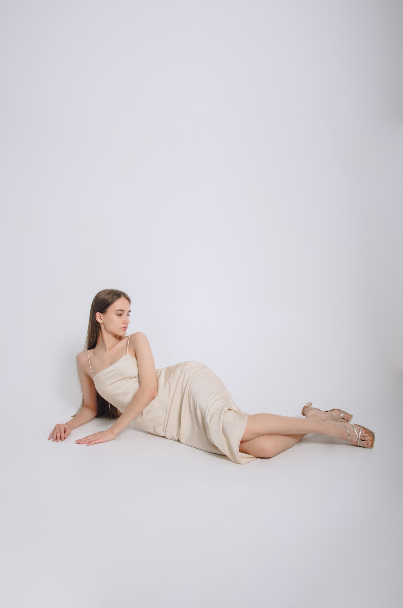 Kobieta w satynowej sukience i złotych sandałach leży na jasnoszarym tle, odwrócona w lewo, z delikatnym makijażem i subtelną biżuterią.