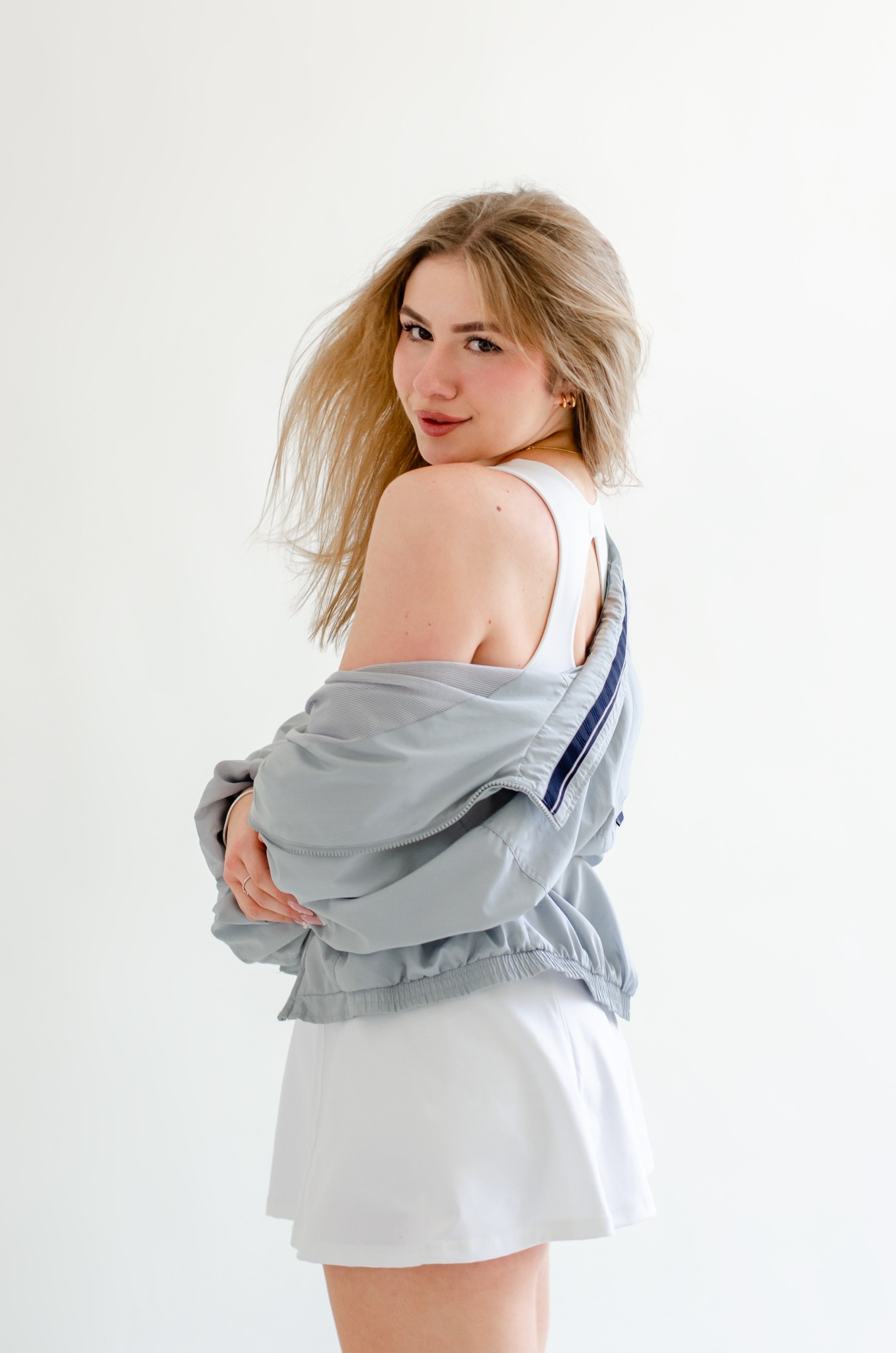 Portret kobiety z blond włosami, odwróconej przez ramię, ubrana w biały top i spódnicę oraz szarą kurtkę sportową na białym tle.